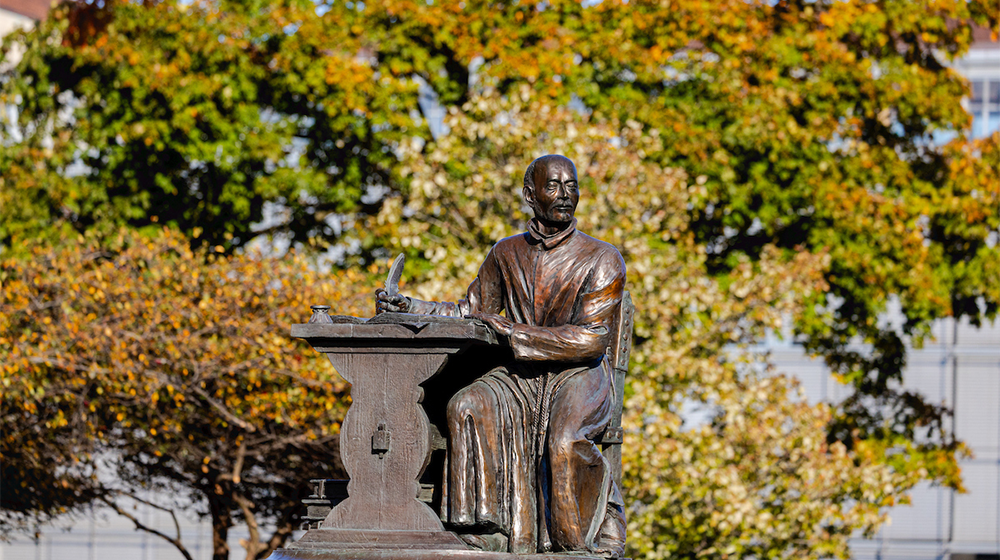 A statue of St. Ignatius writing at his desk in front of fall foliage.